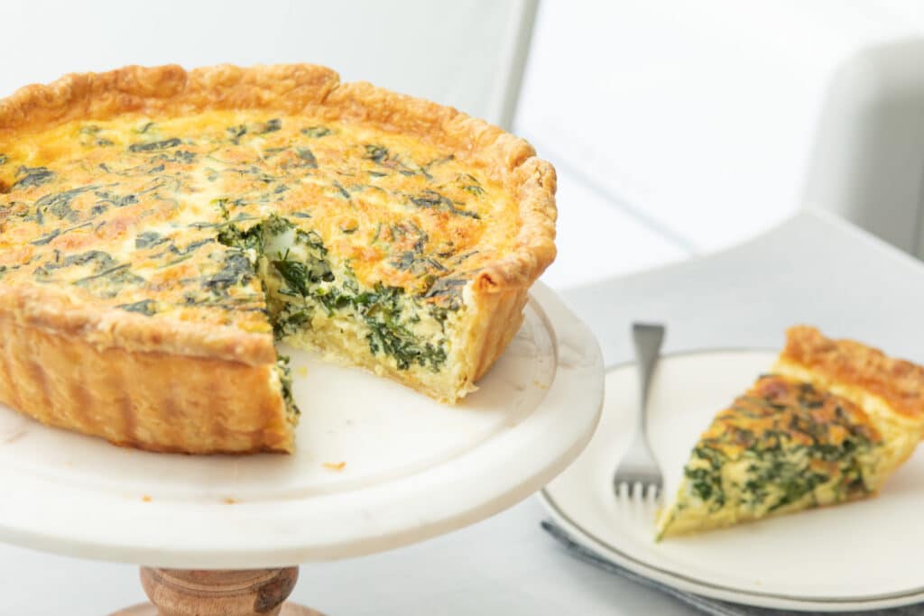 Savory spinach quiche with a golden crust displayed on a marble cake stand, featuring a slice removed and plated beside it, showcasing the creamy filling and vibrant green spinach. Perfect for brunch or a light meal.