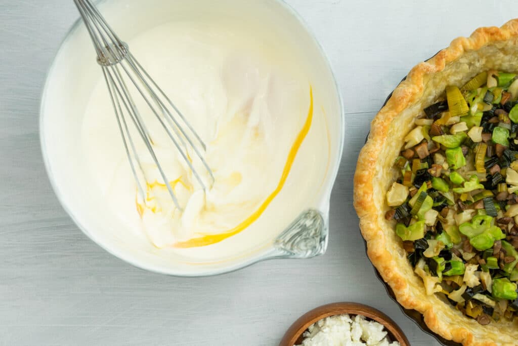 Whisking creamy filling in a bowl next to a savory vegetable pie filled with leeks, mushrooms, and green peppers, accompanied by a small bowl of crumbled cheese.
