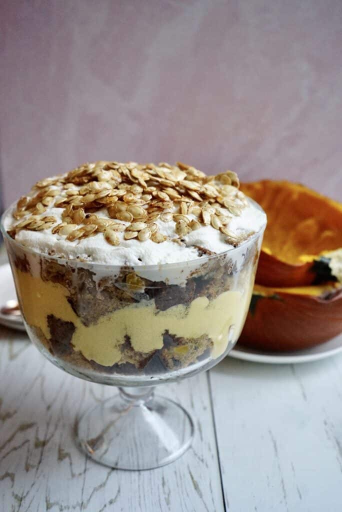 Layered pumpkin trifle featuring sponge cake, creamy custard, and whipped topping, garnished with toasted pumpkin seeds, with a halved pumpkin in the background on a rustic wooden table.