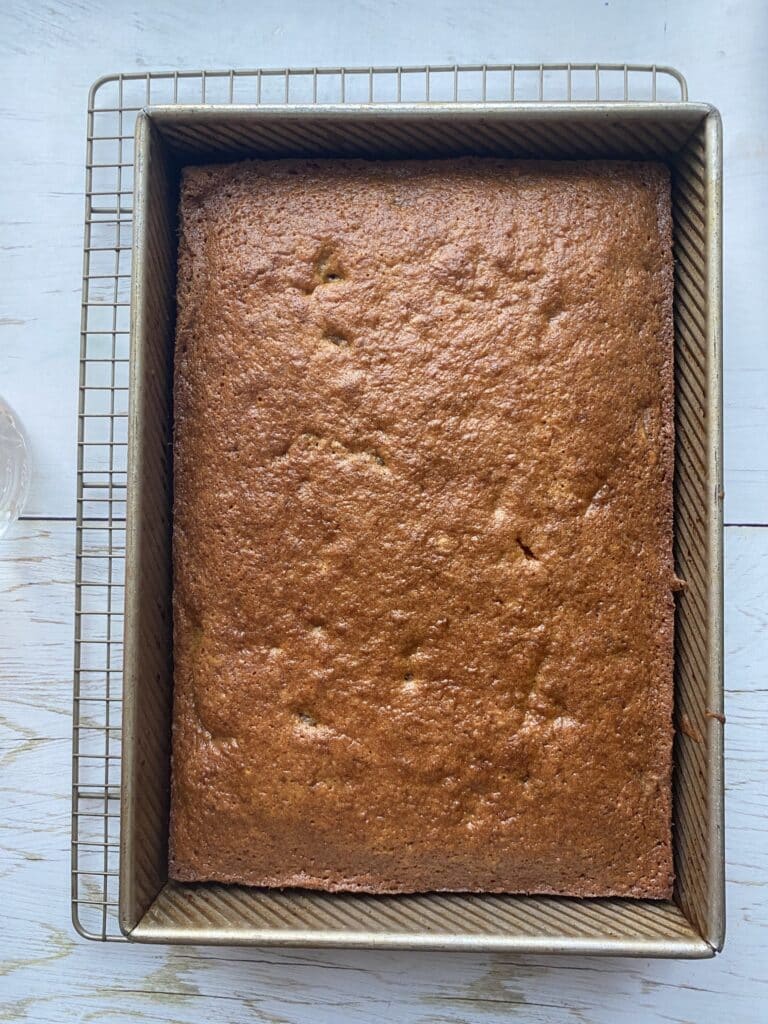 Freshly baked golden brown cake in a rectangular metal baking pan, placed on a cooling rack, with a light wooden background. Ideal for dessert recipes or baking inspiration.