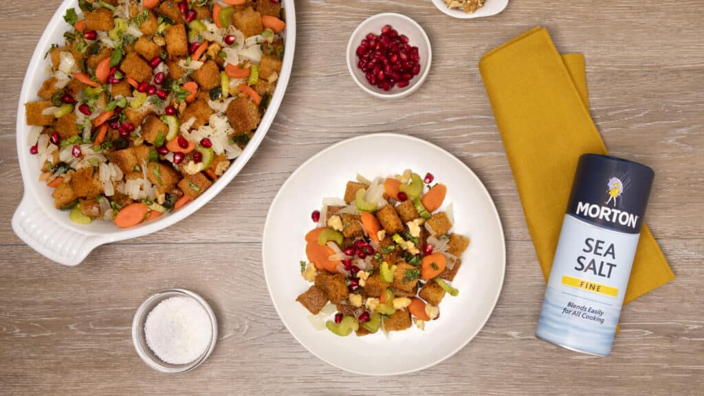 Colorful vegetable stuffing with croutons, garnished with pomegranate seeds and walnuts, served in a white dish alongside a plate of the same dish and Morton Sea Salt on a wooden table.