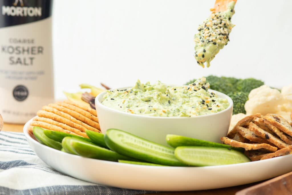 Creamy green dip served in a white bowl, surrounded by fresh cucumber slices, crispy crackers, and assorted vegetables including broccoli and cauliflower, with a container of coarse kosher salt in the background. Perfect for healthy snacking or entertaining guests.