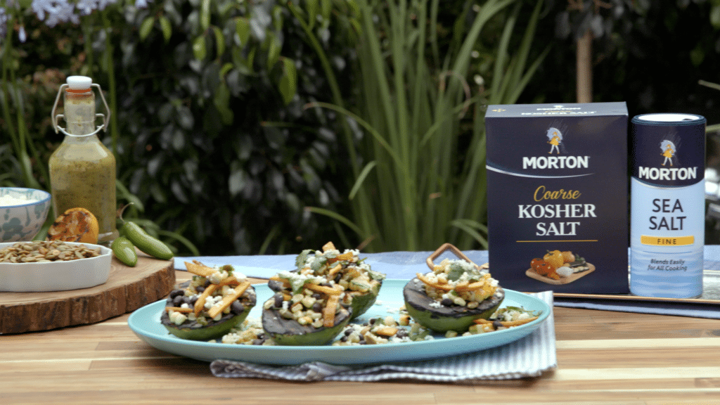 Colorful stuffed avocados topped with black beans, corn, and fried onions, accompanied by Morton Coarse Kosher Salt and Sea Salt. A bottle of dressing and a bowl of pumpkin seeds are also visible, set against a lush green backdrop. Perfect for healthy meal inspiration.