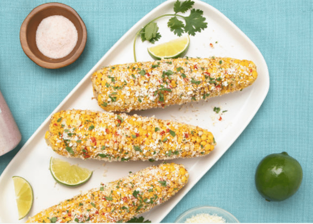 Three flavorful Mexican street corn cobs topped with cheese, cilantro, and chili flakes, served on a white platter with lime wedges and a small bowl of salt, against a turquoise background.
