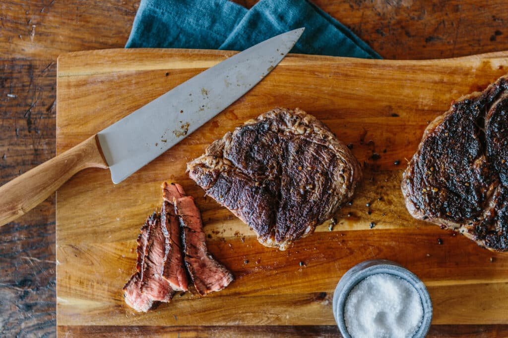 Sliced beef steak on a wooden cutting board with a sharp knife and a small bowl of salt, showcasing the juicy texture and rich color of the meat.