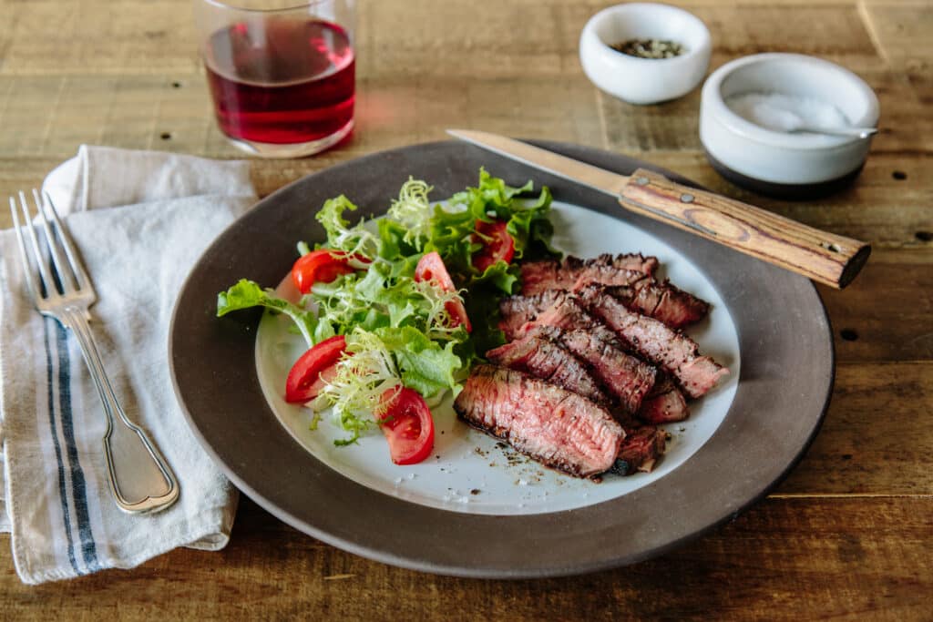 A plate of sliced grilled steak served with a fresh salad of mixed greens and cherry tomatoes, accompanied by a glass of red beverage, salt, and pepper on a rustic wooden table.