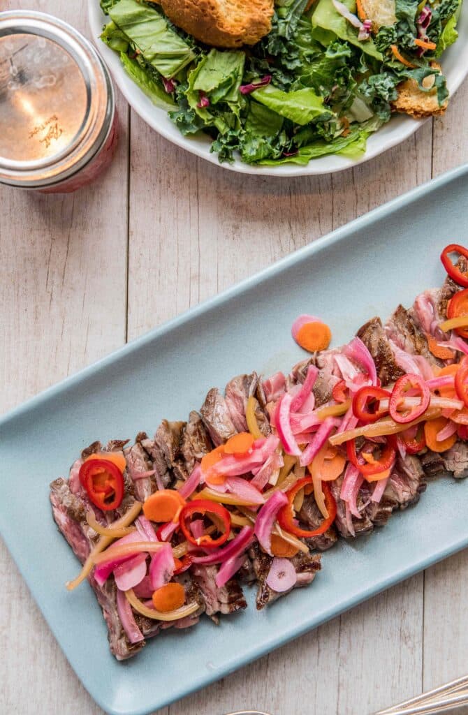 Grilled steak slices topped with colorful pickled onions, carrots, and chili peppers on a light blue platter, accompanied by a fresh green salad and a mason jar on a wooden table.