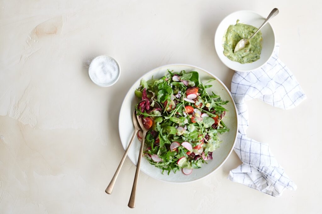 Fresh green salad with mixed greens, cherry tomatoes, and radishes, served in a bowl with two wooden utensils. A small bowl of creamy dressing and a pinch of salt are placed nearby, accompanied by a checkered napkin.