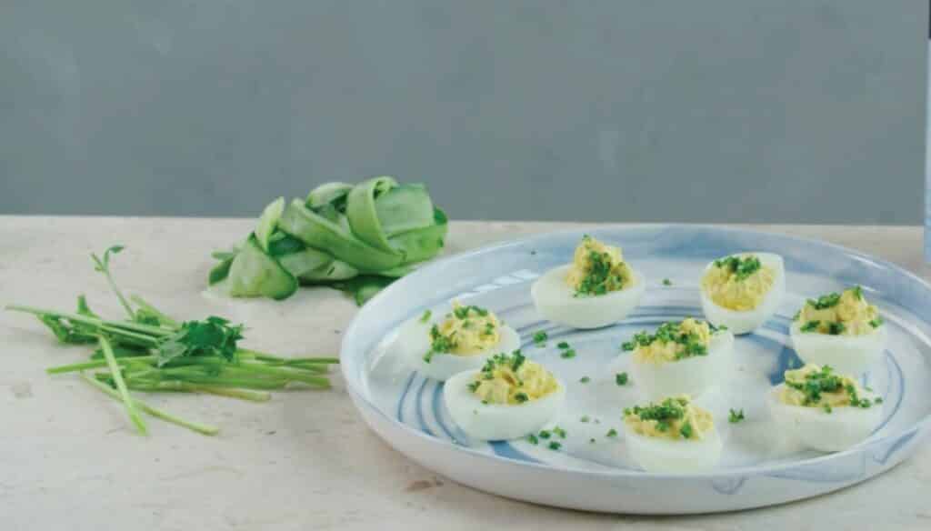 Plate of deviled eggs garnished with fresh herbs, surrounded by cucumber peels and green onions on a marble surface. Perfect appetizer for gatherings and parties.