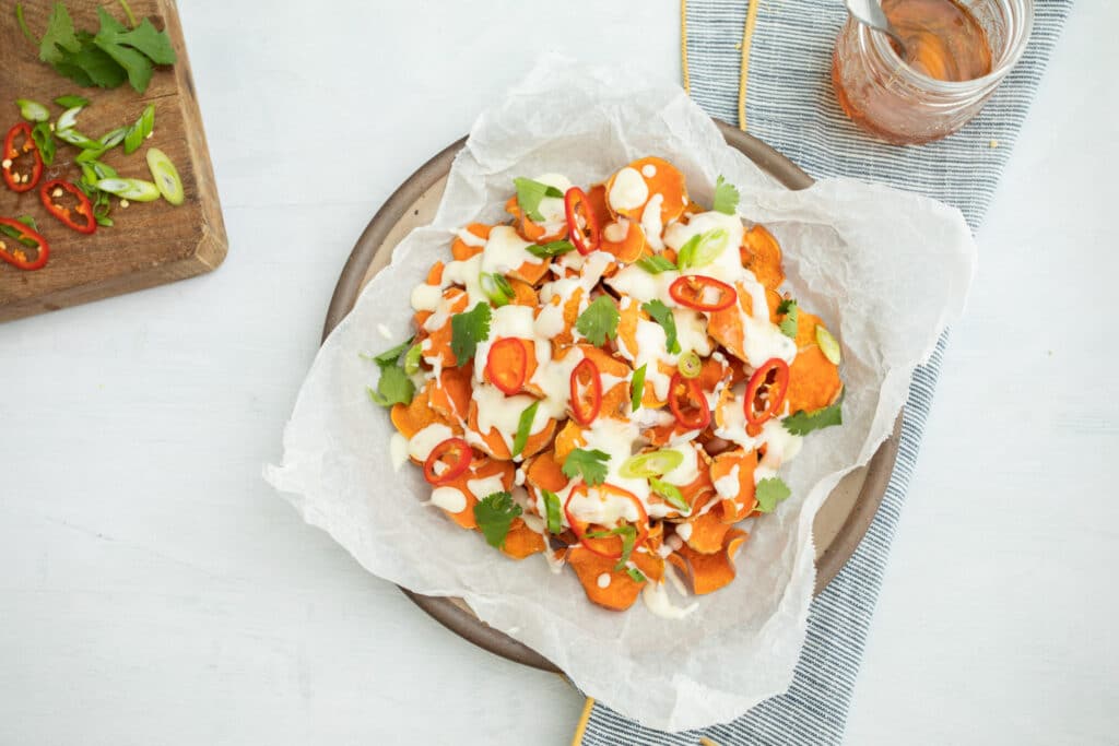 Sweet potato nachos topped with creamy sauce, sliced red chilies, green onions, and fresh cilantro, served on a rustic plate with parchment paper. A jar of sauce and chopped vegetables are in the background, creating a vibrant and appetizing presentation.