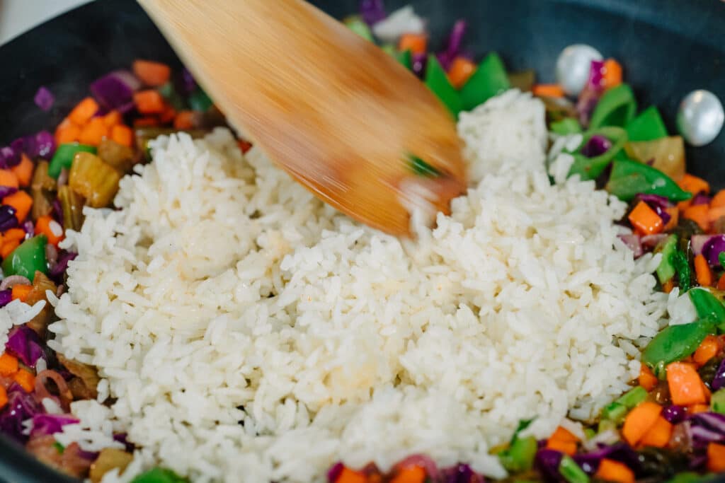 Stirring cooked white rice with colorful mixed vegetables in a frying pan, showcasing a wooden spatula and vibrant ingredients like carrots, green beans, and purple cabbage.