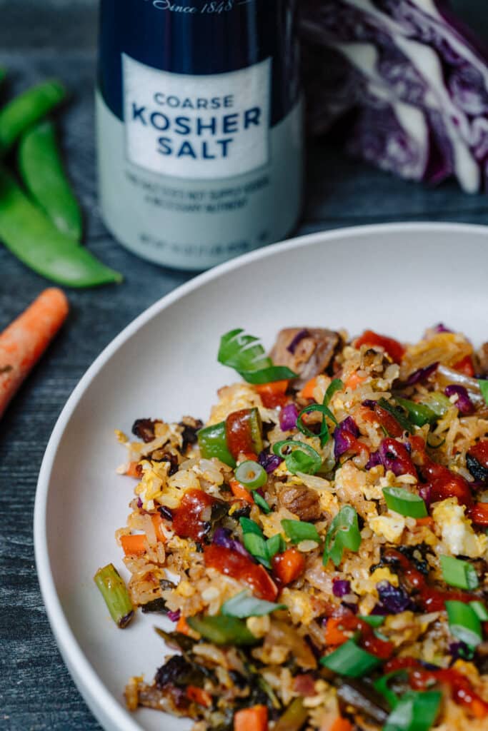 Colorful vegetable fried rice served in a white bowl, featuring vibrant ingredients like bell peppers, carrots, and green onions, with a container of coarse kosher salt in the background.