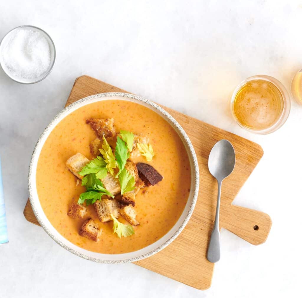 Bowl of creamy tomato soup topped with croutons and fresh celery leaves, served on a wooden cutting board alongside a spoon and a glass of beer, with a small bowl of salt in the background.