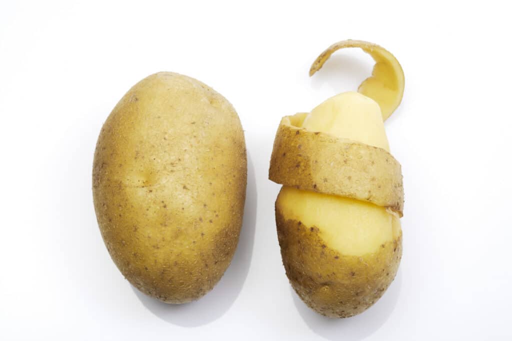 Close-up of a whole potato next to a partially peeled potato with a curling skin, set against a white background. Ideal for illustrating potato preparation or cooking processes.