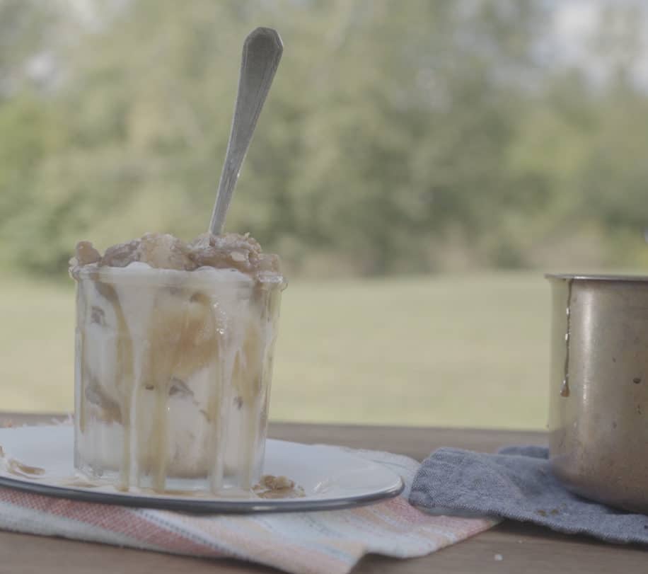 Delicious dessert featuring a glass cup filled with creamy ice cream, topped with caramel sauce and crushed nuts, set against a blurred green outdoor background. A metal spoon rests in the ice cream, while a small pot sits nearby, adding to the rustic ambiance.