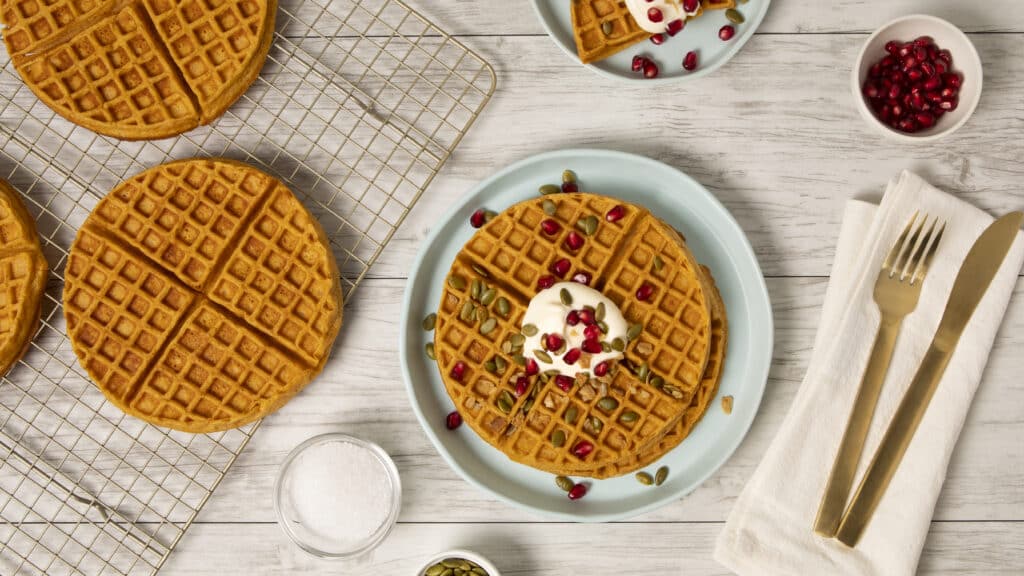 Golden-brown waffles topped with yogurt, pomegranate seeds, and pumpkin seeds on a light blue plate, accompanied by additional waffles on a cooling rack, a small bowl of pomegranate seeds, and a container of sugar, set on a rustic wooden surface.