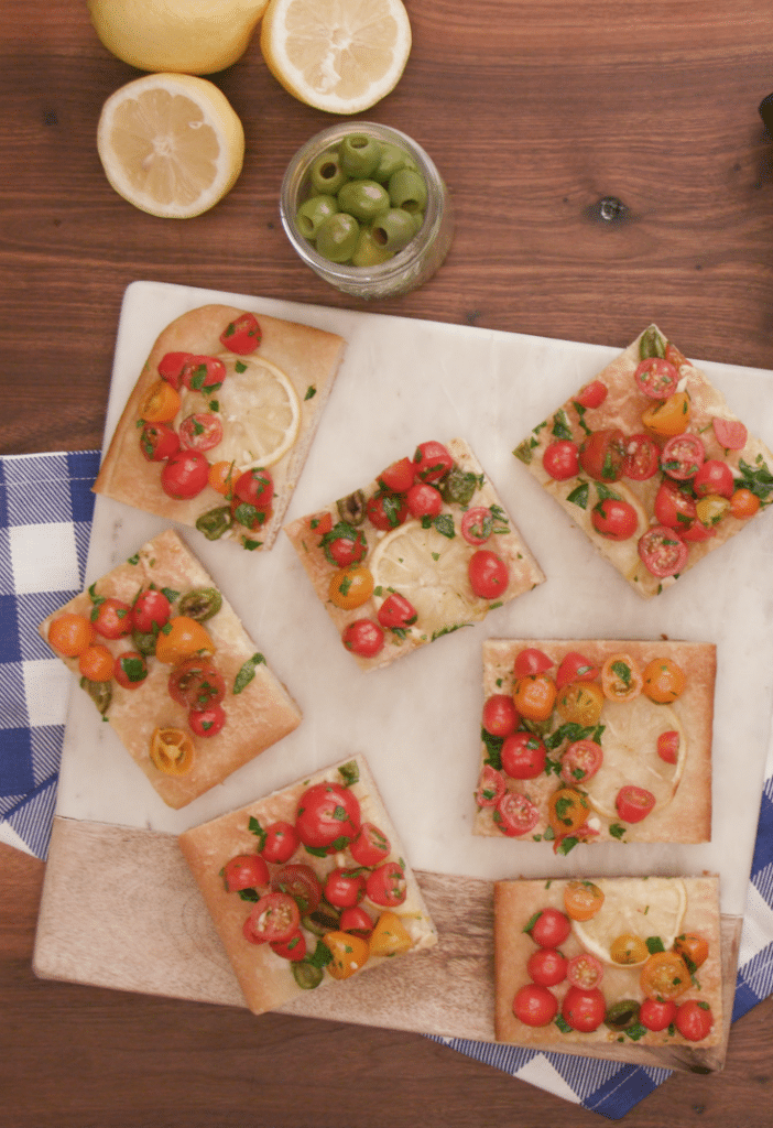 Colorful flatbreads topped with cherry tomatoes, fresh herbs, and lemon slices, arranged on a marble cutting board. Slices of lemon and a jar of green olives are also visible, enhancing the vibrant presentation of this delicious appetizer.