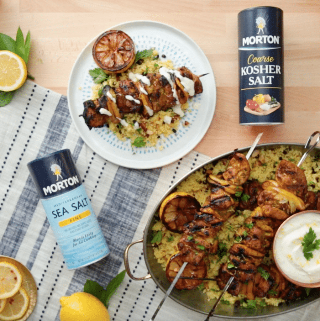 Delicious grilled chicken skewers served over flavorful couscous, garnished with fresh herbs and a side of yogurt dip. Featured Morton Sea Salt and Coarse Kosher Salt for seasoning, with lemon slices enhancing the dish's presentation. Perfect for a summer barbecue or family dinner.