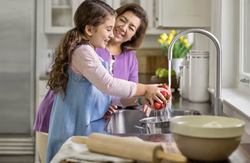 A young girl washes fresh tomatoes in a modern kitchen sink while her smiling grandmother observes, showcasing a warm family cooking moment. Bright yellow tulips and kitchen utensils are visible, highlighting a cheerful cooking atmosphere.