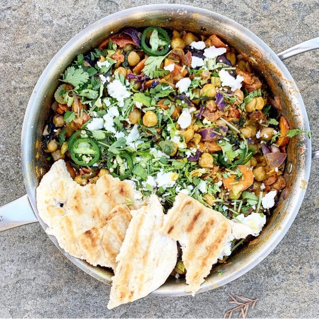 Vibrant chickpea and vegetable stir-fry topped with fresh cilantro, jalapeño slices, and crumbled cheese, served with grilled flatbread in a stainless steel pan.