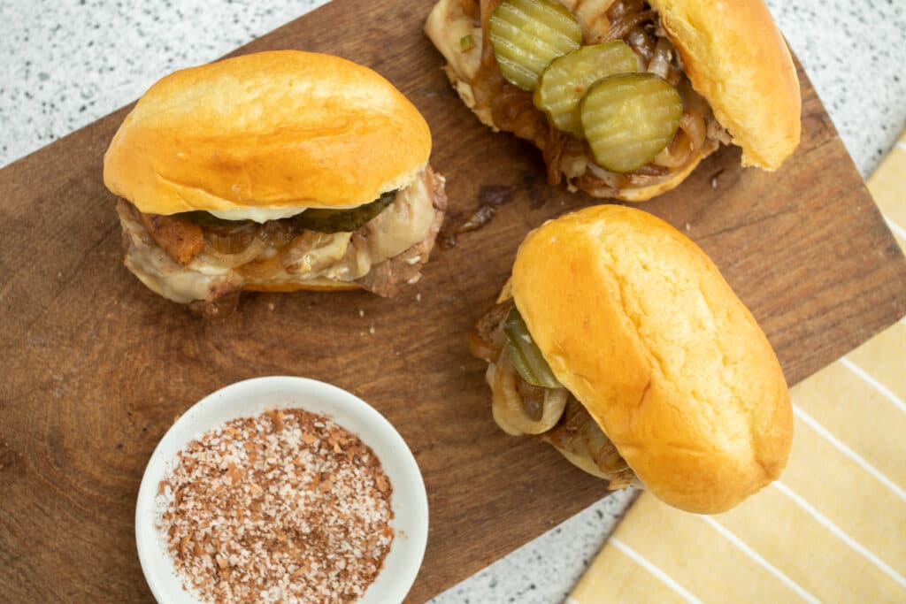 Three delicious sandwiches featuring juicy beef, sautéed onions, and pickles on soft buns, served on a wooden cutting board alongside a small bowl of seasoning.