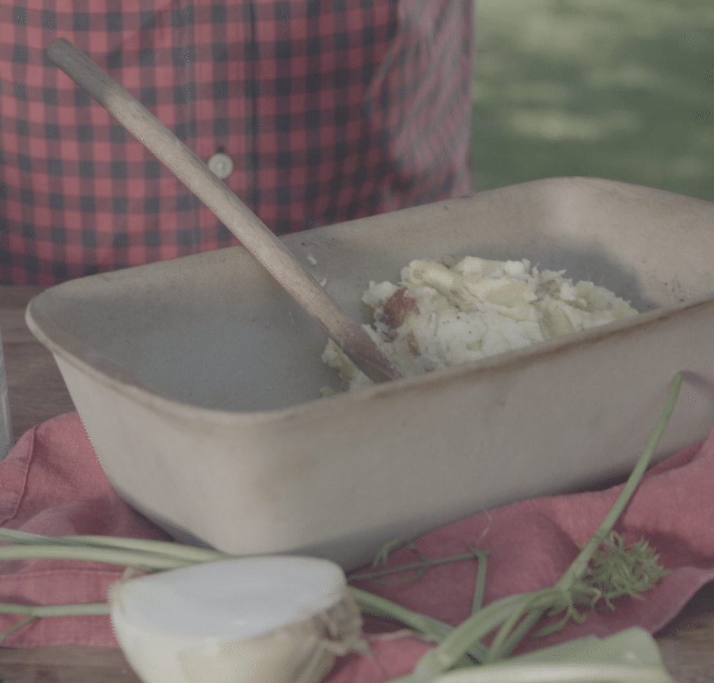 Bowl of creamy mashed potatoes with a wooden spoon, placed on a rustic table with fresh vegetables and a red cloth, capturing a cozy outdoor dining experience.