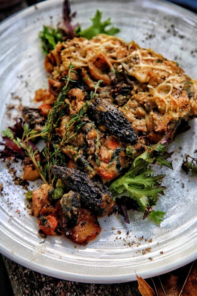 A delicious serving of baked dish featuring morel mushrooms, mixed vegetables, and herbs, presented on a textured white plate with a bed of fresh greens.
