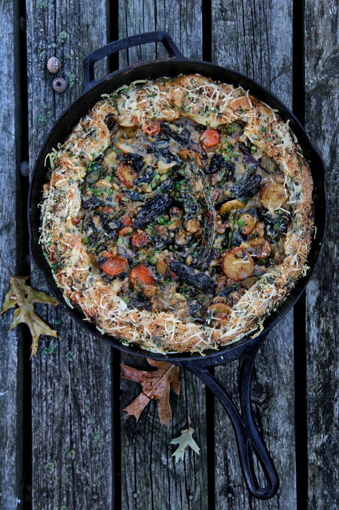 Savory baked dish in a cast iron skillet, featuring a mix of sautéed vegetables, herbs, and melted cheese, set on rustic wooden planks with autumn leaves. Perfect for a cozy meal or gathering.