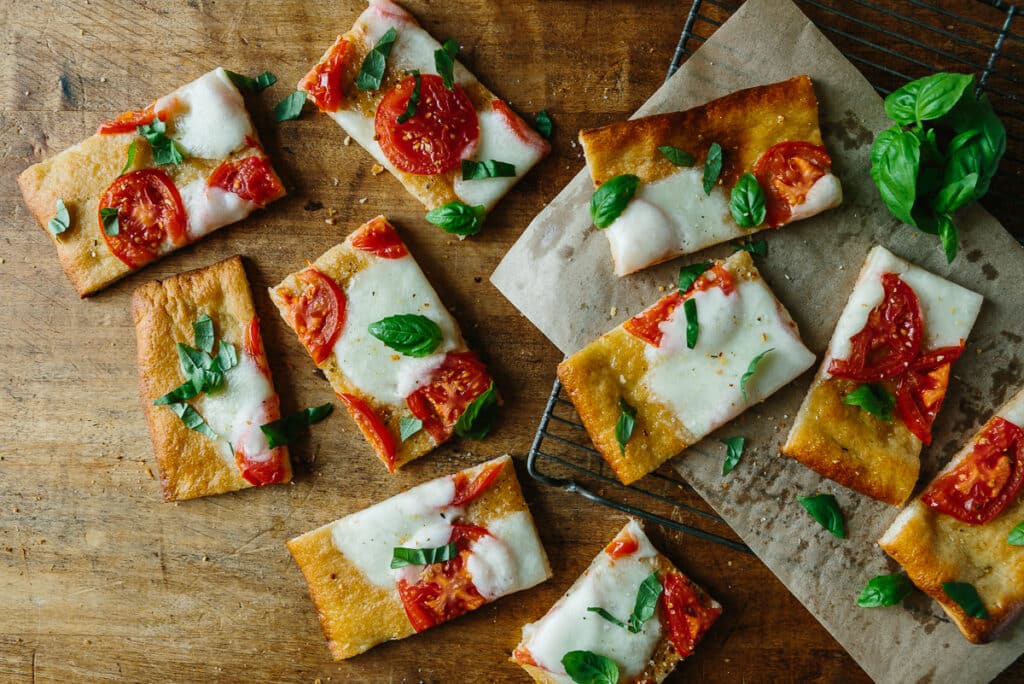 Freshly baked rectangular flatbreads topped with mozzarella cheese, sliced tomatoes, and fresh basil leaves, arranged on a wooden surface and parchment paper. Perfect for a delicious appetizer or snack option.