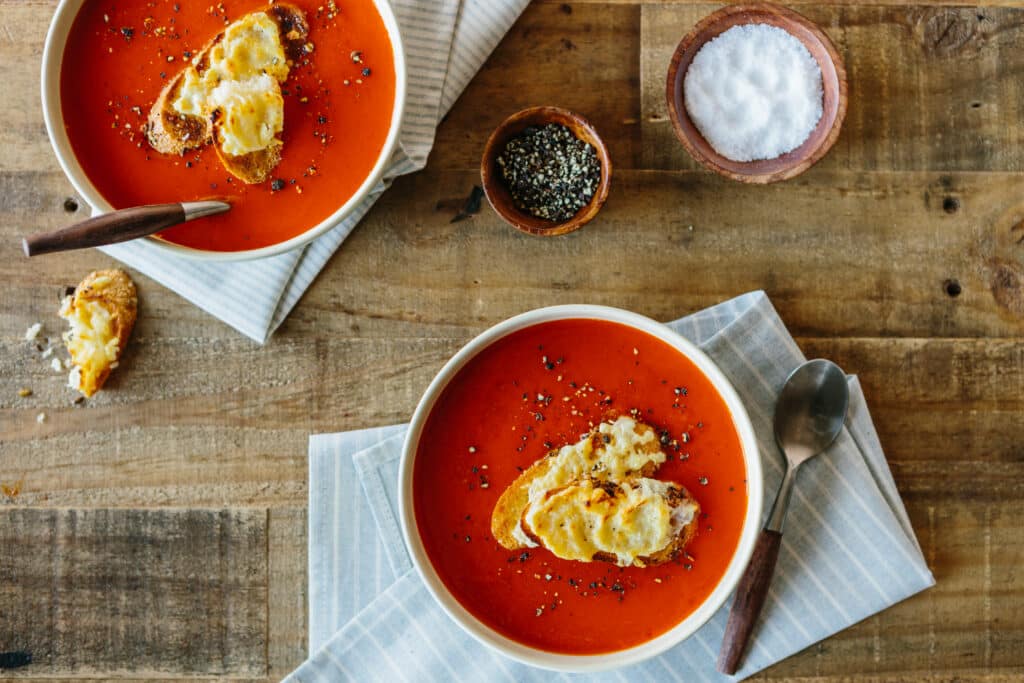 Creamy tomato soup served in white bowls with toasted bread topped with melted cheese, garnished with black pepper, and accompanied by salt and mixed herbs on a rustic wooden table.