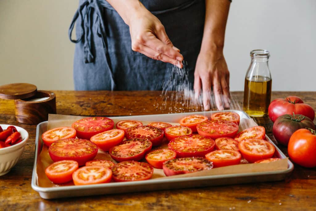A person in a gray apron sprinkles salt over sliced tomatoes arranged on a baking sheet, with a bowl of whole tomatoes and a bottle of olive oil nearby, on a wooden countertop.
