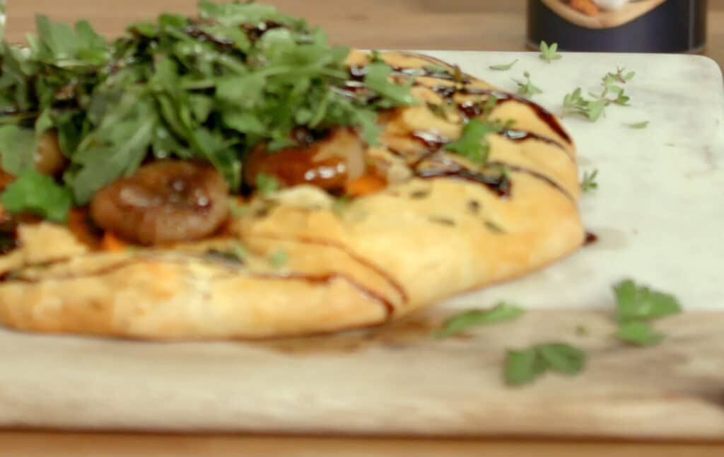 Savory flatbread topped with fresh arugula, roasted vegetables, and a drizzle of balsamic glaze, served on a wooden cutting board.