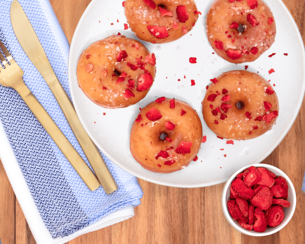 Glazed donuts topped with crushed strawberries on a white plate, accompanied by a bowl of fresh raspberries and gold cutlery on a wooden table.