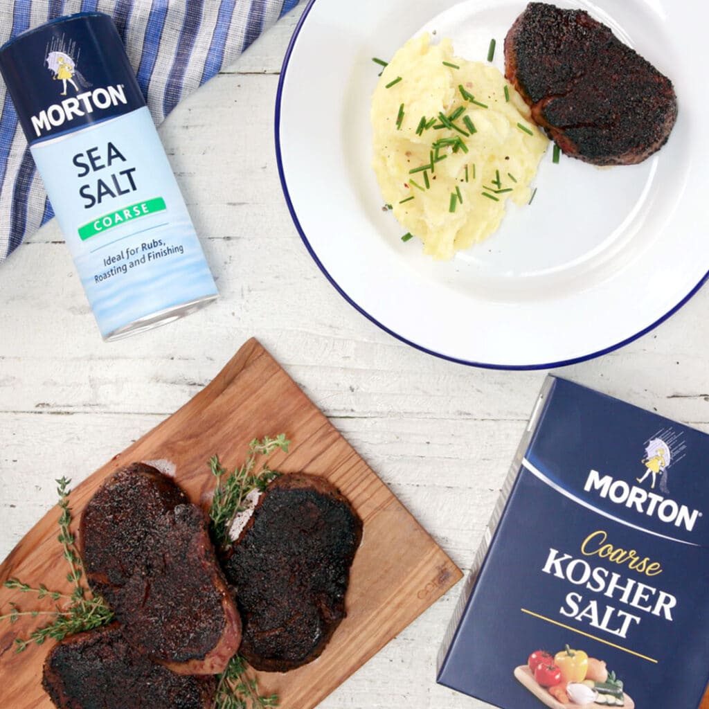 Image of a delicious meal featuring seasoned steaks on a wooden cutting board, accompanied by creamy mashed potatoes garnished with chives on a white plate. A can of Morton Sea Salt and a box of Morton Coarse Kosher Salt are visible, highlighting seasoning options for enhancing flavor in cooking.