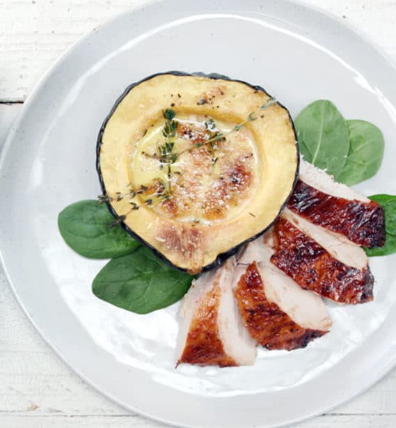 Grilled chicken breast slices served on a plate with fresh spinach leaves and a roasted acorn squash half topped with herbs and seasoning.
