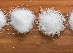 Variety of salt types displayed on a wooden surface, showcasing different textures and grain sizes, including fine salt and coarse salt.