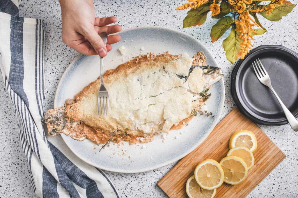 A hand holding a fork is poised over a baked whole fish garnished with herbs and a crispy coating, served on a light blue plate; lemon slices and a black plate are nearby, with a striped towel and decorative flowers in the background.