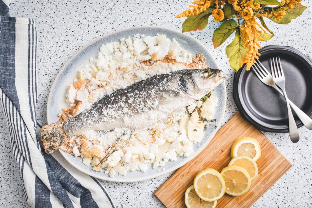 Whole fish coated in salt on a serving platter, surrounded by fresh herbs, with lemon slices on a wooden cutting board and utensils nearby, set against a speckled countertop.
