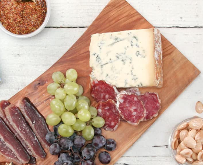 Charcuterie board featuring a variety of gourmet foods, including blue cheese, slices of salami, green and black grapes, and a bowl of mixed nuts, complemented by a container of mustard. Ideal for entertaining or casual dining.
