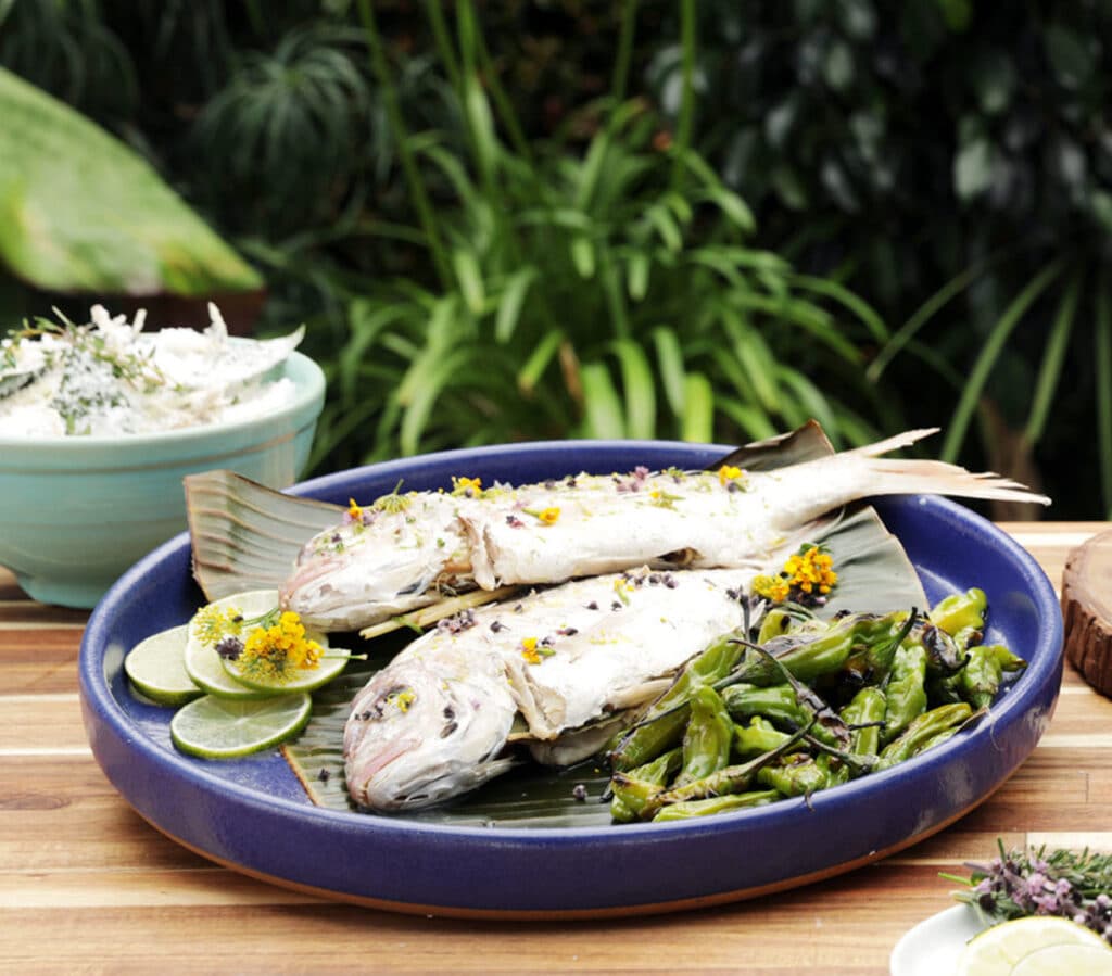 Grilled whole fish garnished with edible flowers, served on a banana leaf, accompanied by lime slices and sautéed green peppers, set against a lush green background.