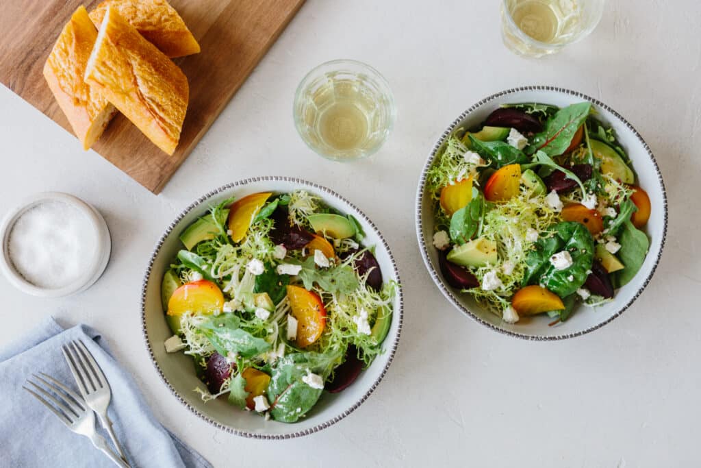 Fresh garden salad featuring mixed greens, colorful heirloom tomatoes, avocado, and feta cheese, served in two bowls alongside crusty bread and a small dish of salt, with glasses of white wine in the background.