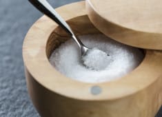 Close-up of a wooden salt cellar with a spoon, containing coarse white salt, placed on a textured gray surface.