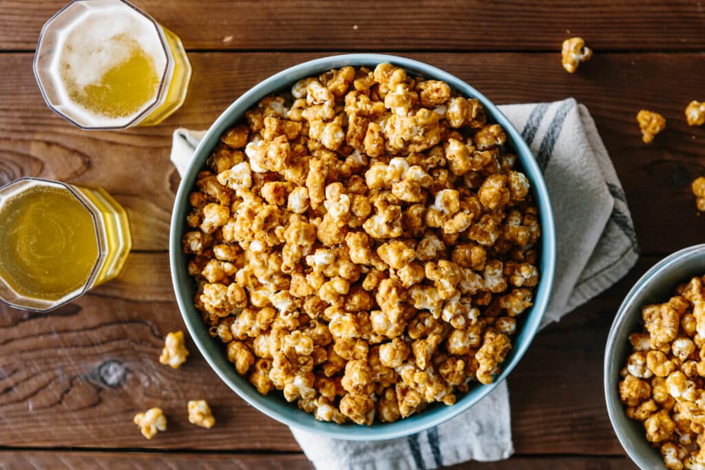 Bowl of caramel popcorn surrounded by glasses of beer on a wooden table, perfect for a movie night or snack time.