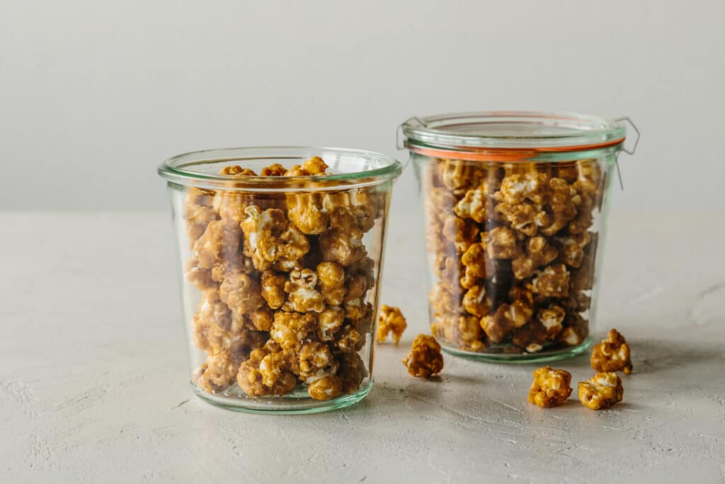 Two glass jars filled with caramel popcorn, placed on a light-colored surface, with a few pieces scattered nearby.