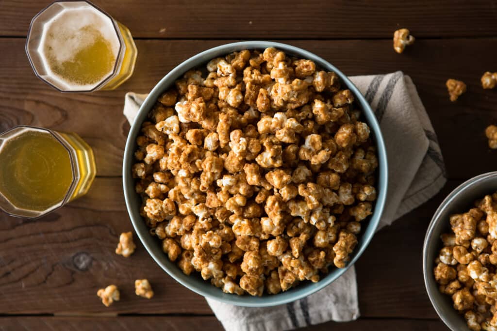 A large bowl of caramel popcorn sits on a wooden table, accompanied by two glasses of golden beer. The scene captures a cozy snack setup, perfect for movie nights or gatherings.