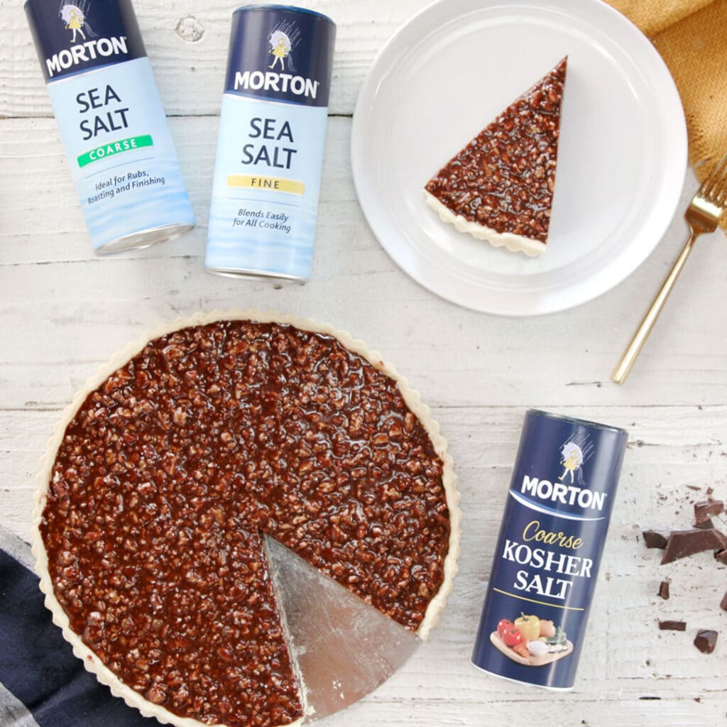 Morton Sea Salt containers alongside a chocolate tart with a slice removed, presented on a rustic wooden surface. The image highlights both coarse and fine sea salt options, emphasizing their culinary uses for enhancing flavors in cooking and baking.