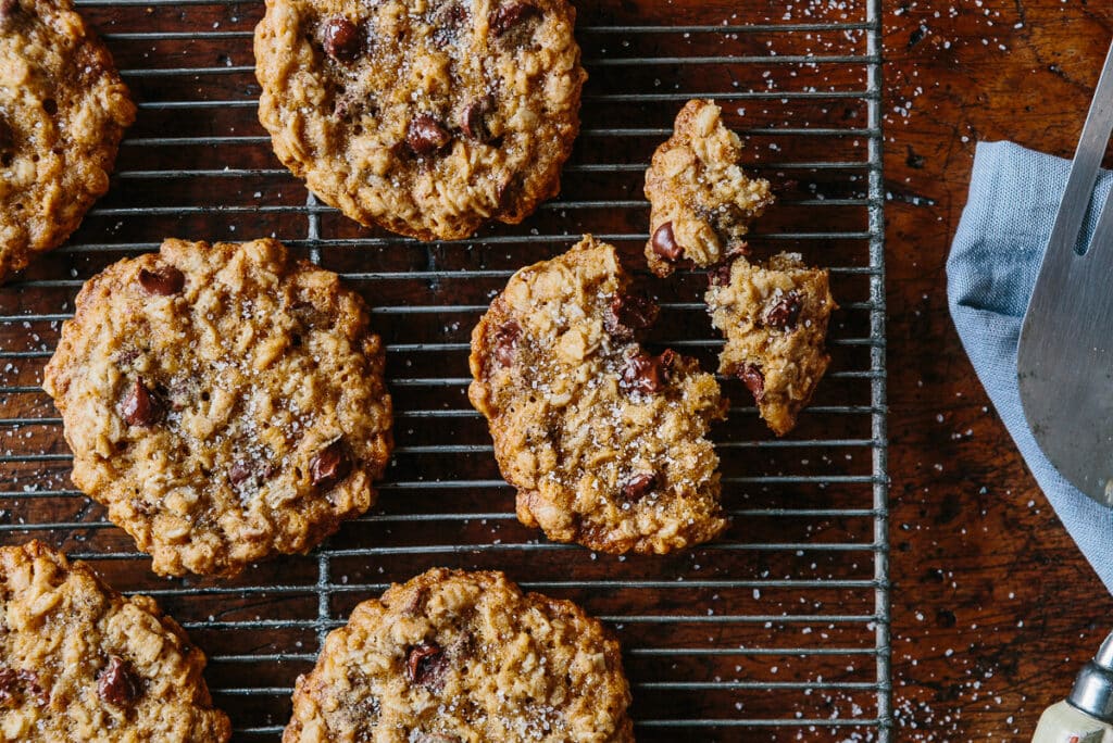 Freshly baked oatmeal chocolate chip cookies cooling on a wire rack, with a few cookies broken apart, showcasing their chewy texture and sprinkled with sea salt.