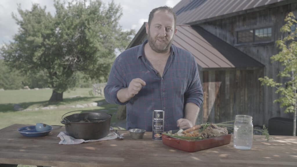 Chef demonstrating outdoor cooking techniques with a cast iron pot, fresh vegetables, and kosher salt on a wooden table, set against a rustic backdrop of trees and a barn.