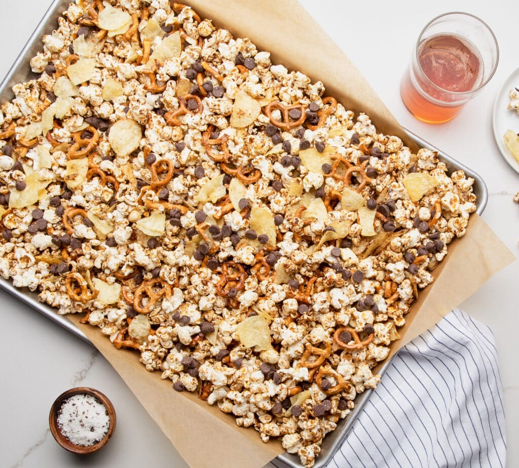 Tray of sweet and salty popcorn mix topped with pretzels, potato chips, and chocolate chips, served with a glass of beverage. Ideal for snacks and party treats.