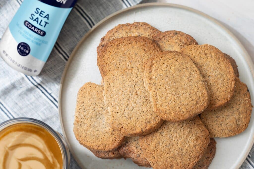 Plate of freshly baked, golden-brown cookies with a coarse sea salt container and a jar of peanut butter in the background, showcasing a delicious snack idea.